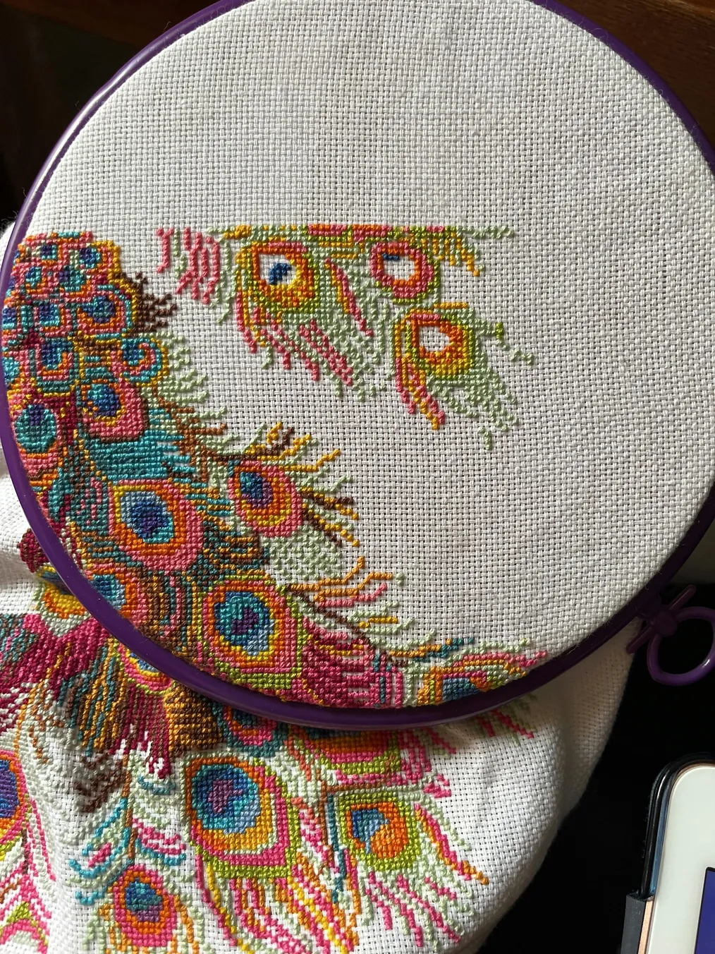 Colorful peacock feather design in embroidery hoop, showcasing intricate cross-stitching artistry.